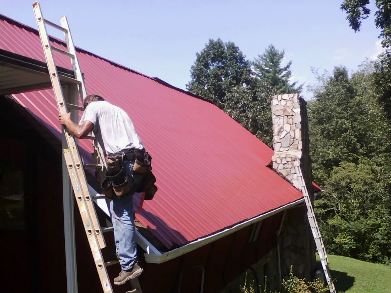 Roofer on a ladder beside a red metal roof and stone chimney — Soffit & Fascia Repair by Roofing Brothers in Belle Center